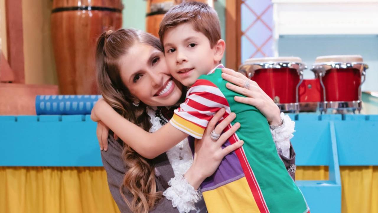 Juliana D'Agostini sorri abraçando o filho de seis anos, que veste camiseta nas cores amarelo, vermelho, verde e roxo. Ao fundo, tambores e instrumentos.