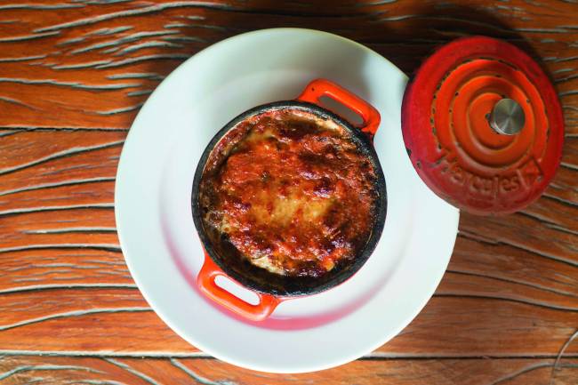 Parmigiana di melanzane_8_Johnny Mazzilli.jpg (1) Panelinha de ferro vermelha contendo um parmigiana apoiada sobre um prato raso branco em uma mesa de madeira