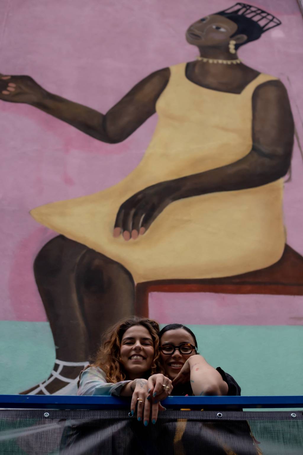 Larissa de Souza e Pam Araújo posam lado a lado em frente a grande mural com pintura de mulher negra sentada com vestido amarelo.