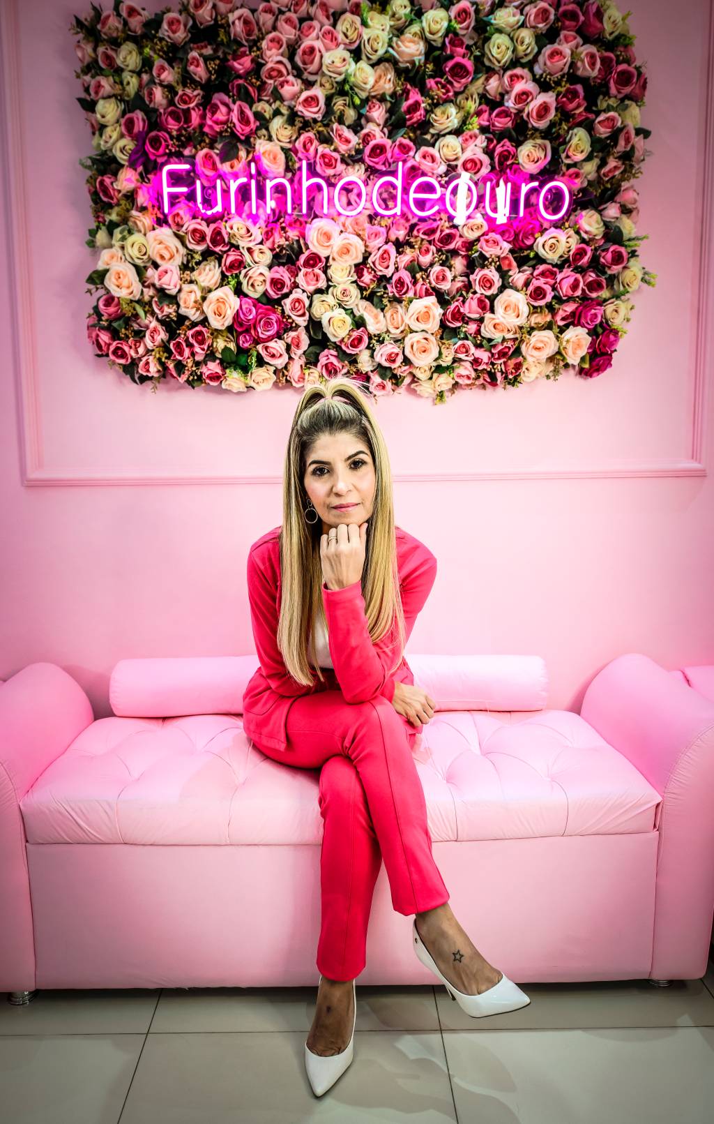 Rachel Strazzeri, da Furinho de Ouro, posa de pernas cruzadas em sofá rosa claro. Veste blazer e calça rosa, em tom mais escuro, apoia uma das mãos no queixo e encara a câmera. Ao fundo, na parede, um letreiro em neon com flores exibe o título Furinho de Ouro.