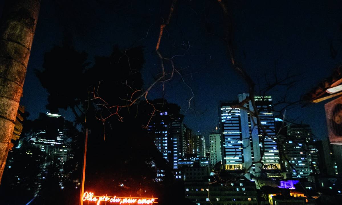 Espaço externo com letreiro neon vermelho onde se lê olha pro céu meu amor, com pessoas reunidas conversando