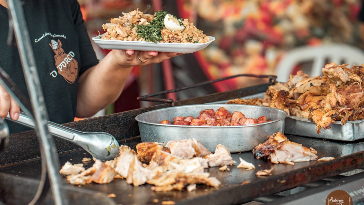 Foto de pessoa grelhando carne em uma chapa e segurando bandeija com alimentos.