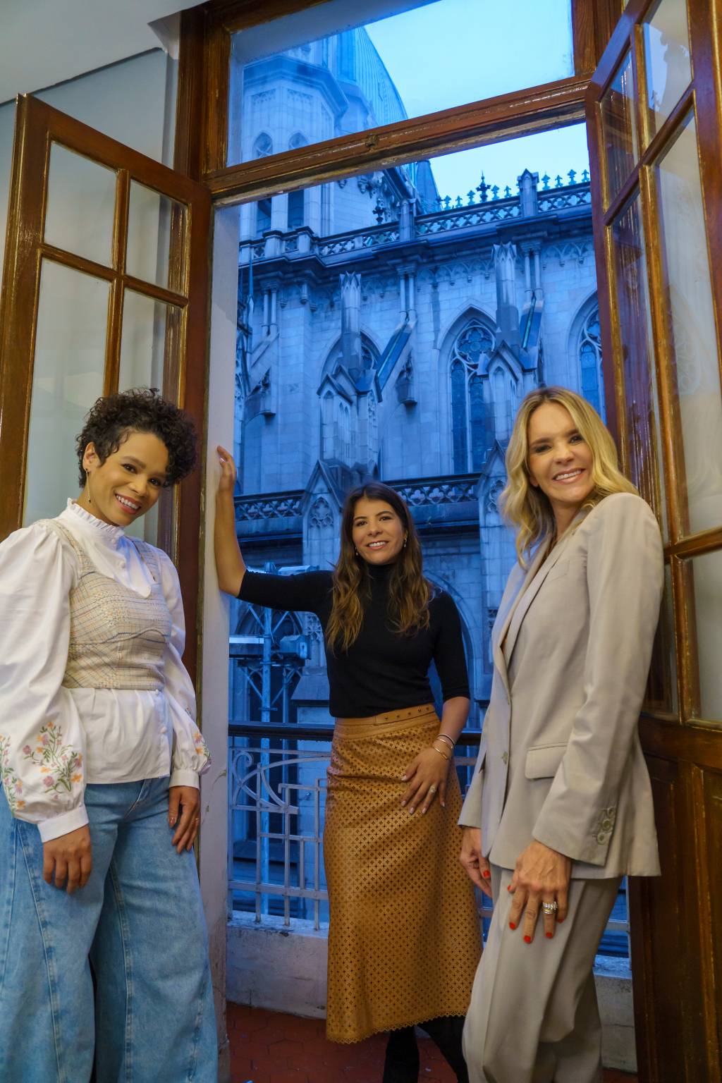 Tatiane, Bianca e Deborah (a partir da esquerda) posam de pé olhando para baixo na câmera. As três estão de pé perto de uma janela com vista para a Catedral da Sé.