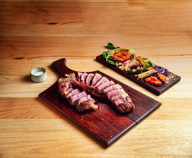 Carne de T-bone fatiada sobre tábua ao lado de tábua com legumes grelhaos