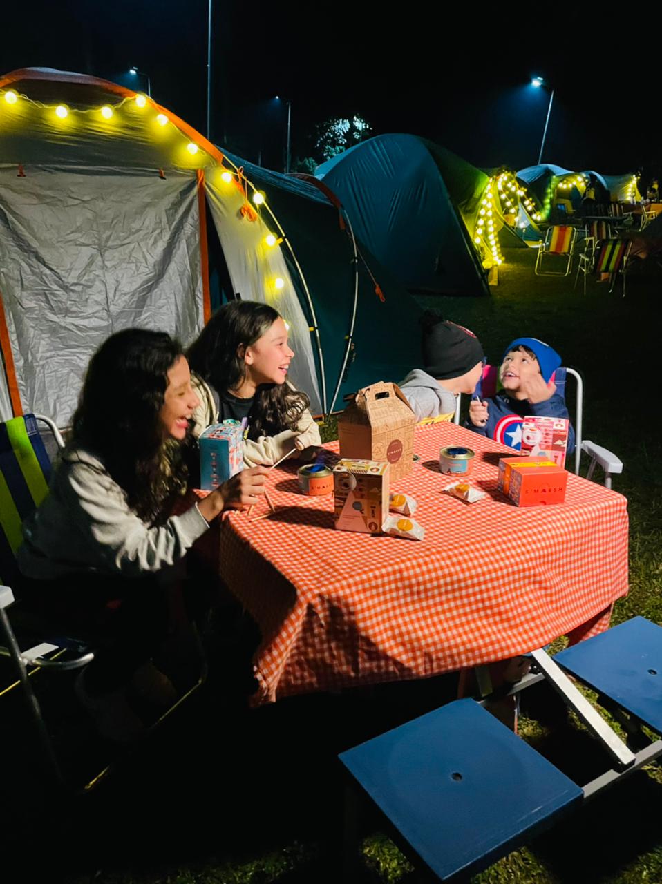 Crianças sentadas em uma mesa de piquenique jantando. Elas estão acampando no Parque Burle Marx à noite e sorriem