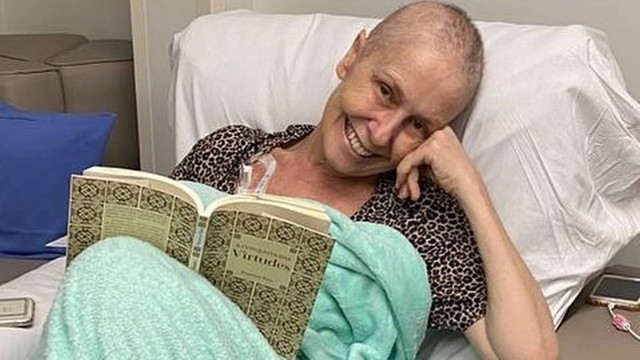 Foto de Susana Naspolini em cama de hospital, sorrindo e segurando livro