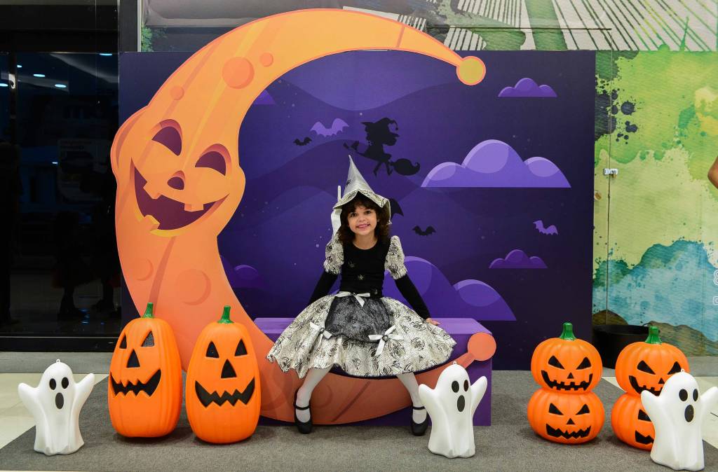 Espaço temático de Halloween com bonecos de abóbora e fantasmas. Menina fantasiada de bruxa está sentada no centro