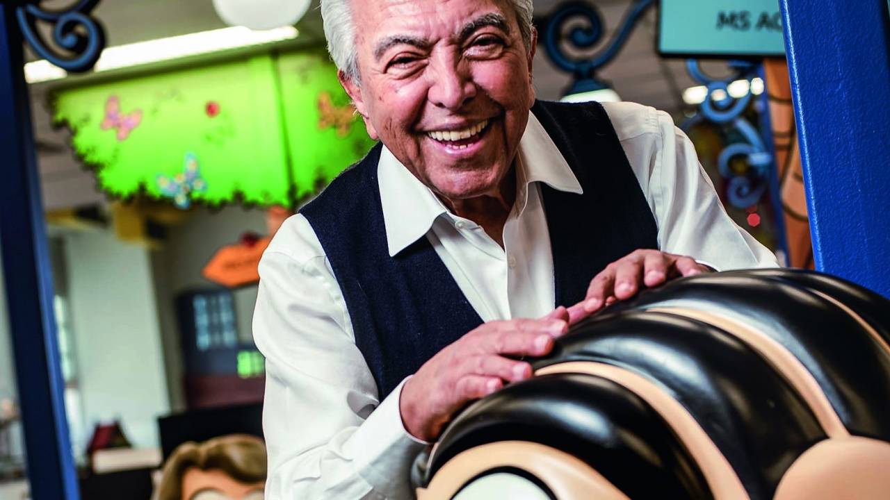Mauricio de Sousa posa sorrindo no estúdio da Turma da Mônica, pousando as mãos sobre boneco da Mônica