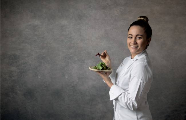 Giovanna Grossi do Animus de pé à frente de uma parede cinza segurando um prato e um garfo e vestindo branco