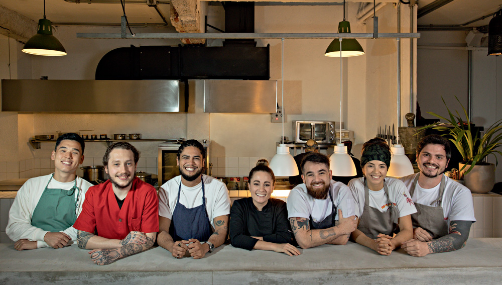 Imagem mostra sete chefs sorrindo, apoiados sobre balcão