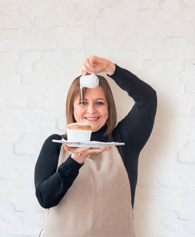 Carla Pernambuco: – Carlota Chef Carla com um prato na mão contendo um suflê sobre o qual despeja o conteudo de um minibule de porcelana, creme de catupiry