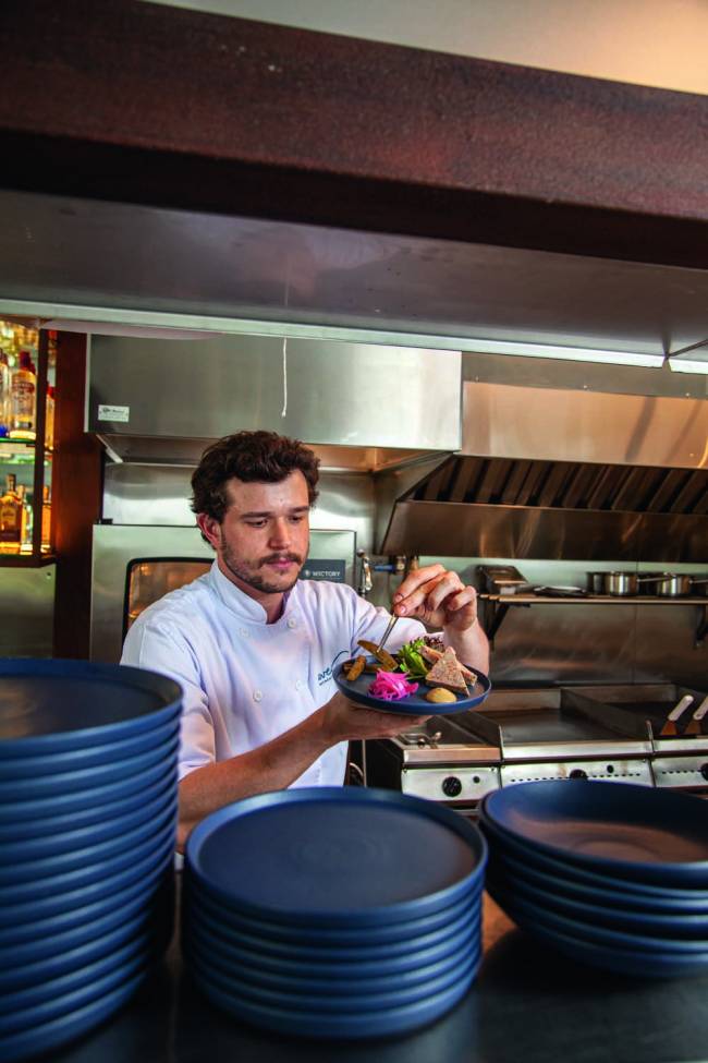 Homem em uma cozinha com revestimento de inox de jaleco branco finaliza um prato em um recipiente de louça azul