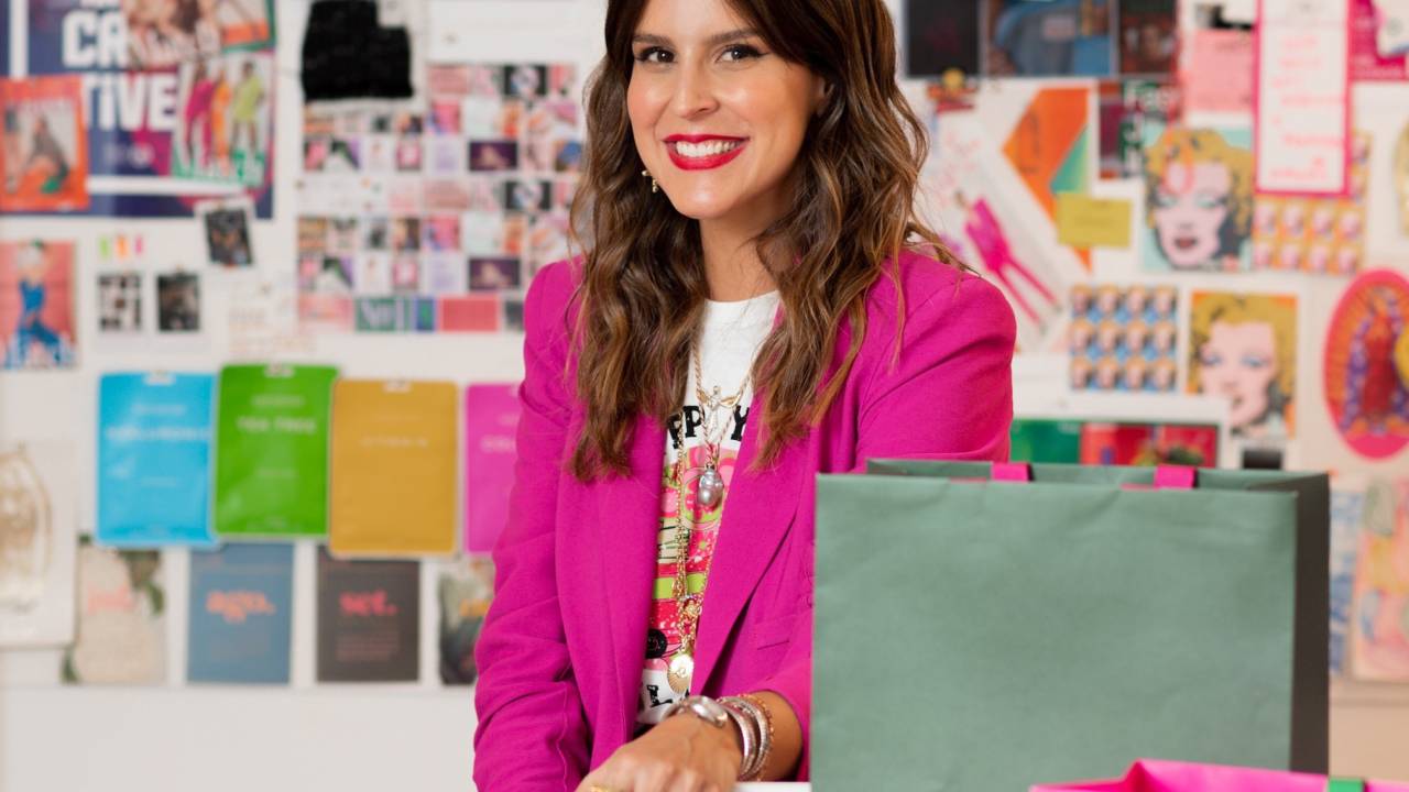 Ciccy Halpern posa sorrindo com blazer rosa pink e sacolas em primeiro plano. Ao fundo, mural repleto de papéis coloridos.