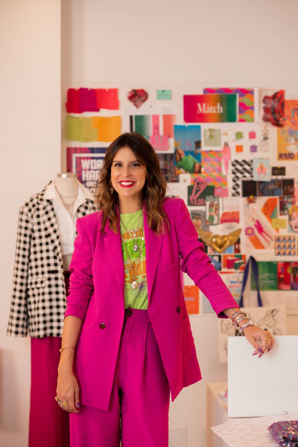 Ciccy Halpern posa sorrindo com blazer rosa pink e sacolas em primeiro plano. Ao fundo, mural repleto de papéis coloridos e um blazer quadriculado em preto e branco.