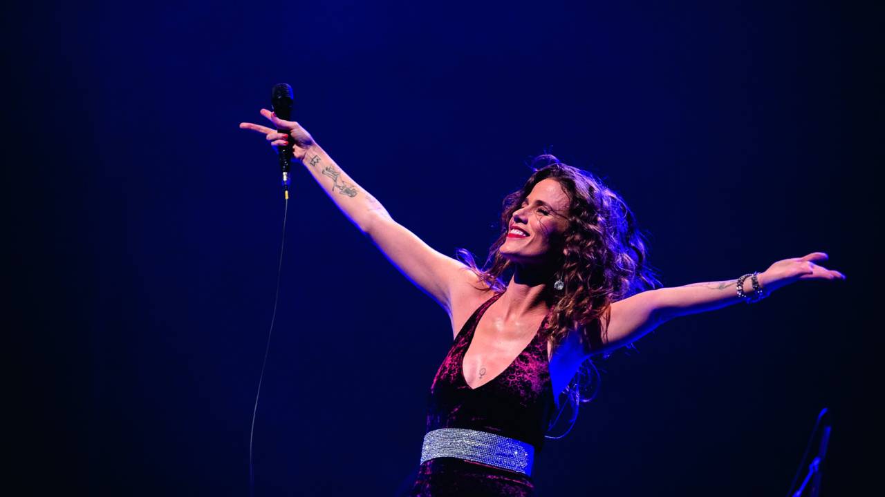 Imagem mostra mulher com microfone na mão e os braços abertos, sorrindo