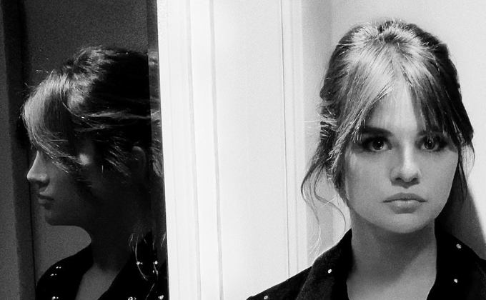 Imagem em preto e branco mostra mulher olhando para frente, à direita, e seu reflexo à esquerda, em um espelho,