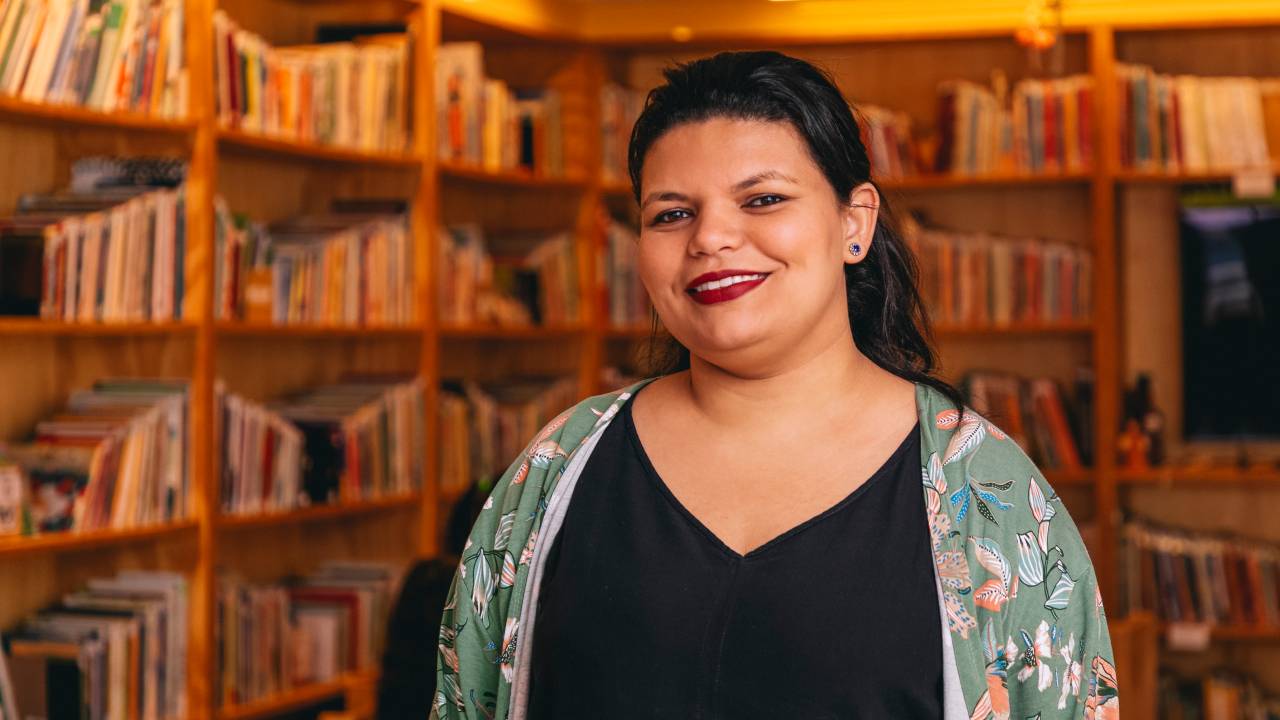 Luana Andrade, pedagoga do Pró-Saber, sorri para a câmera em biblioteca. Veste camiseta preta com blusinha florida.