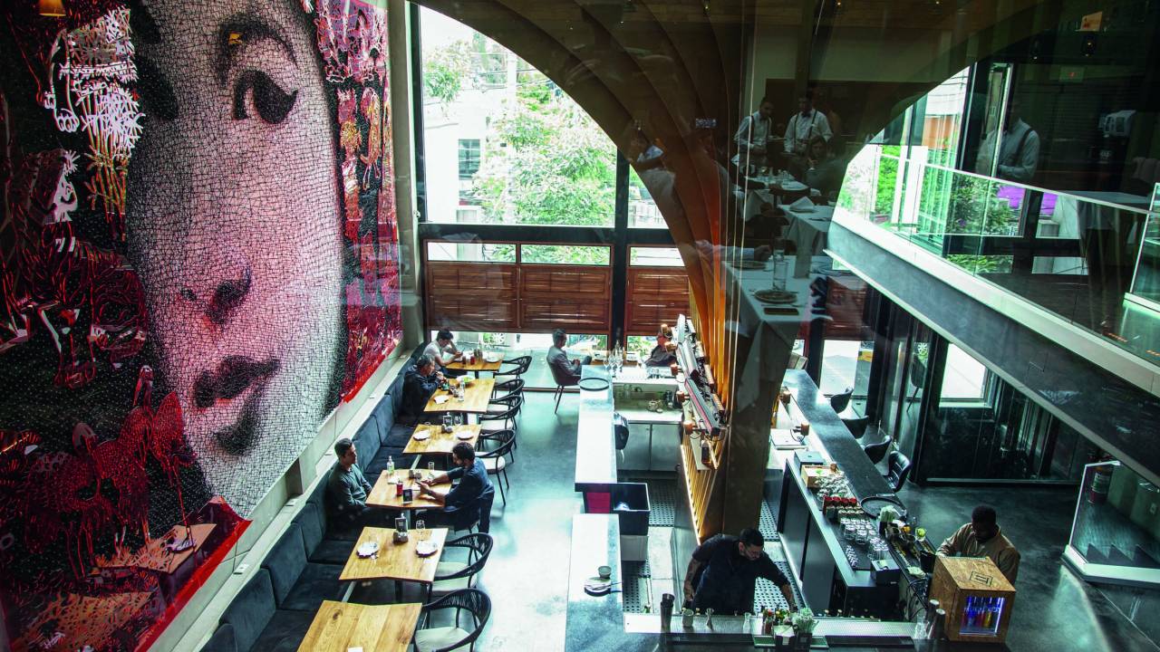 Visão superior de ambiente interno do restaurante, com mural de mulher asiática à esquerda, pilastra e mesas e janelões de vidro ao fundo
