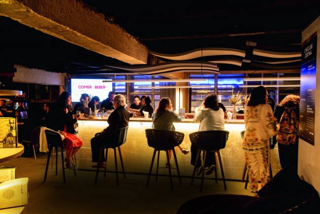 Cineclube Cortina – Feira dos Campeoes Pessoas sentadas em banquetas almofadadas altas em frente a balcão de bar com luz sob o tampo