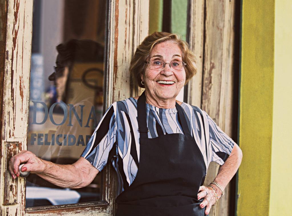 Imagem mostra mulher idosa de avental preto, sorrindo, em frente a restaurante