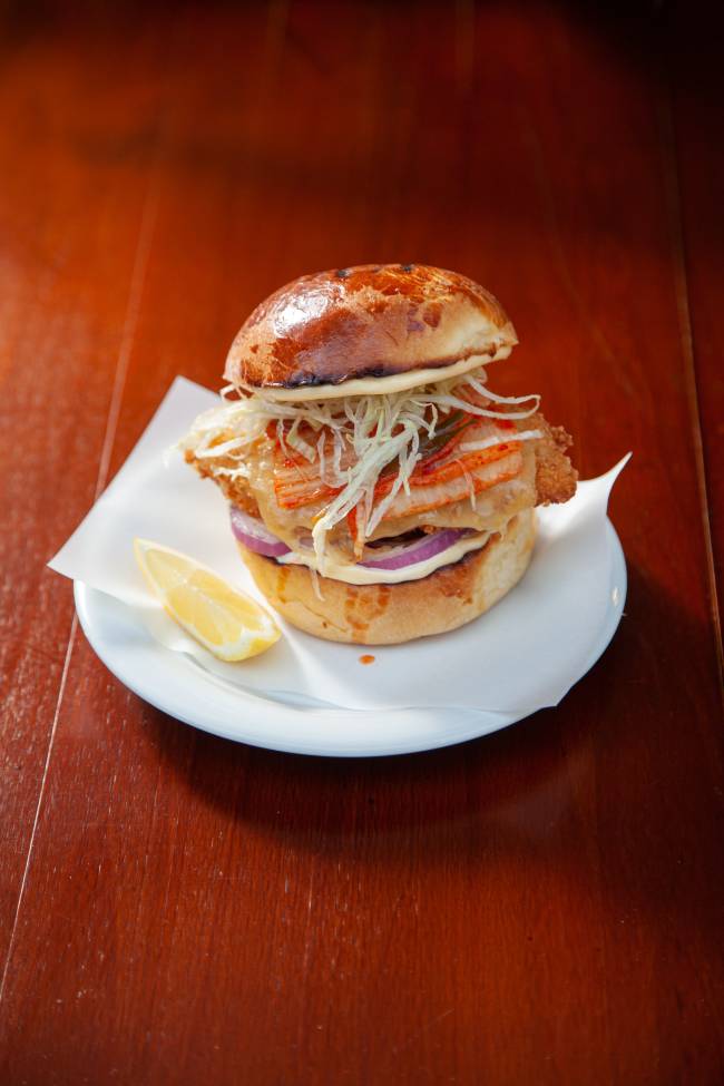 Prato de porcelana sobre mesa de madeira contendo sanduíche de peixe empanado no pão de brioche e fatia de limão