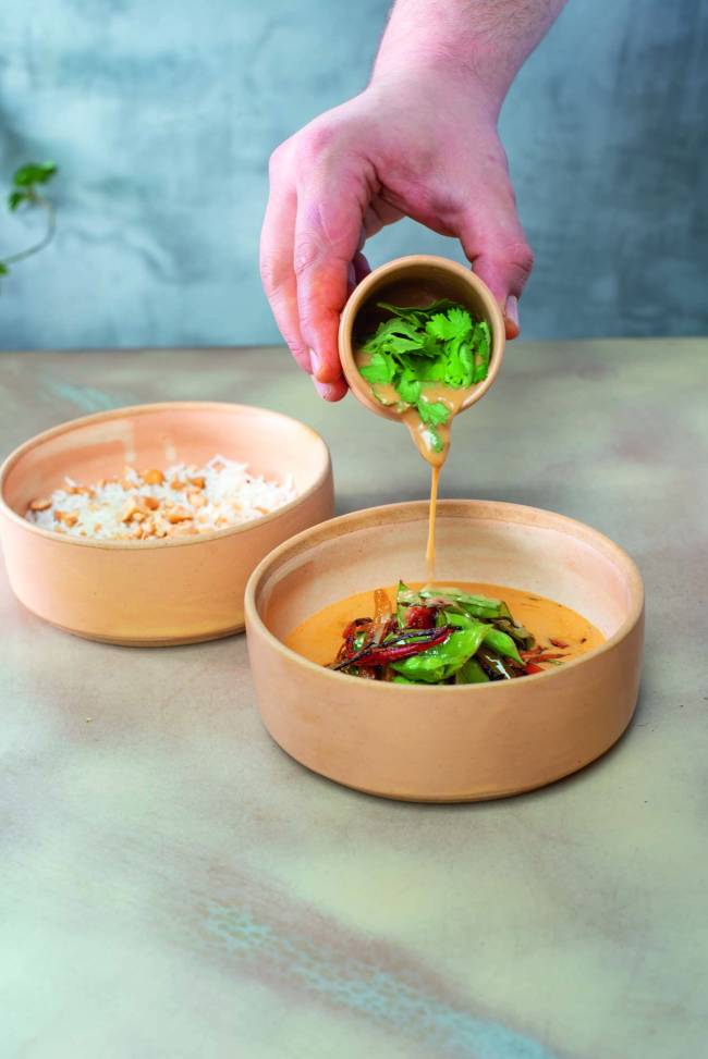 Dois recipientes de cerâmica, um contendo arroz de jasmim e castanha de caju, e o outro um curry de vegetais, no qual uma mão segurando um recipiente despeja curry