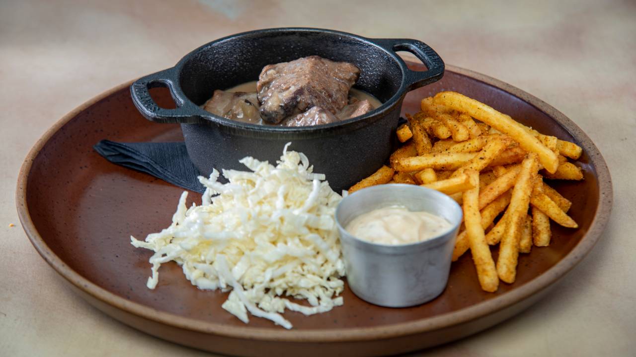 Prato de cerâmica contendo uma panelinha de ferro com carne cozida, acelga cortada em tiras, batatas fritas e molho em um recipiente de alumínio