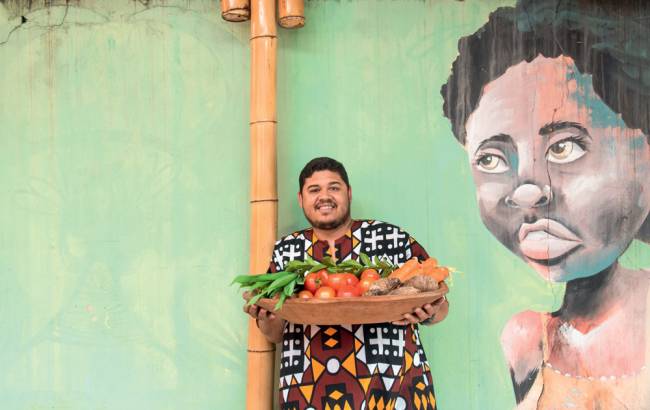 Thiago Vinícius à frente de uma parede na qual há um grafitti de uma mulher negra de blusa laranja, usa uma camiseta de padrão colorido, segura uma cesta de legumes e sorri