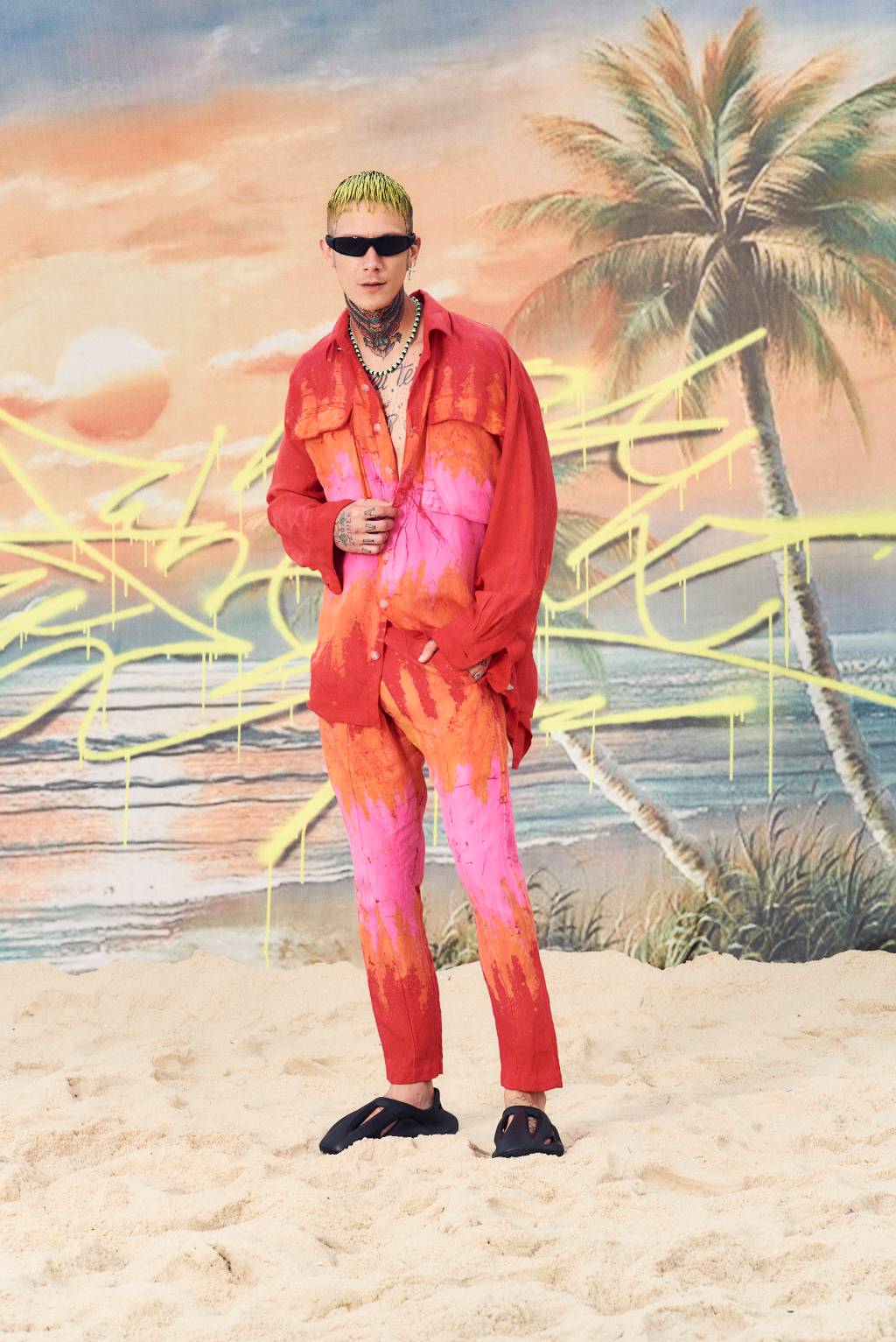Modelo masculino posa de pé com jaqueta e calça alaranjadas, com tons de rosa. Usa óculos escuros, tem cabelos loiros e ao fundo aparece um papel de parede com palmeiras.