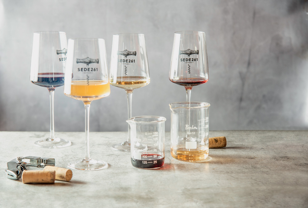 Taças e frascos de vidro de laboratório com vinhos, um diferente do outro. Estão dispostos em uma mesa cinza