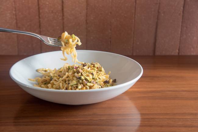 Prato de porcelana branca sobre mesa de madeira contendo espaguete ao pistache com manteiga, aliche italiano e pistaches tostados, e um garfo saindo da esquerda da foto contendo uma porção da massa