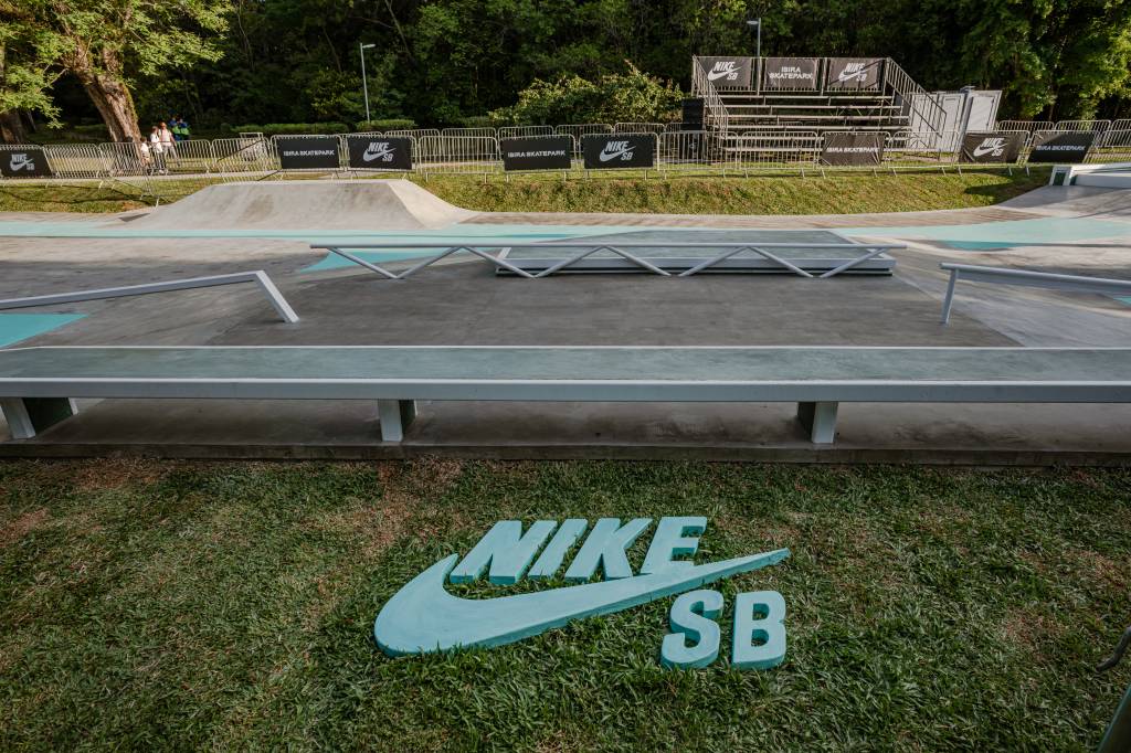 Foto de skate park com faixas azuladas, arquibancada e árvores ao fundo.