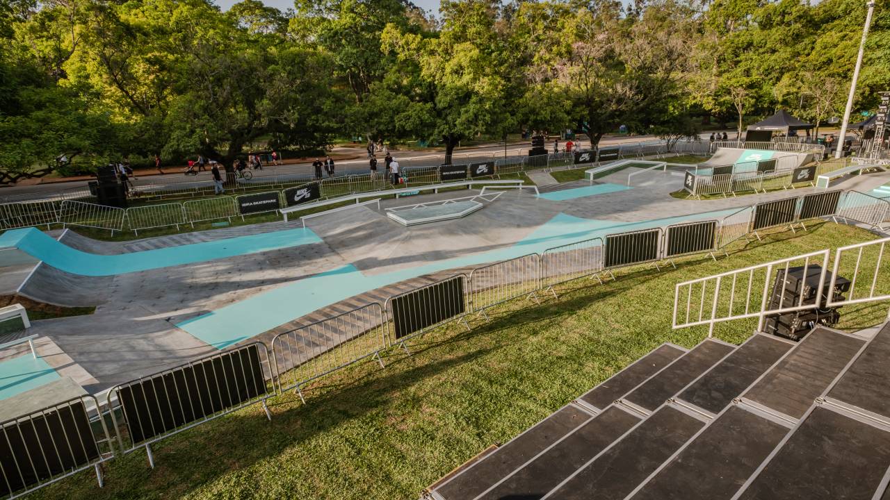 Foto de skate park com faixas azuladas, arquibancada e árvores ao fundo.