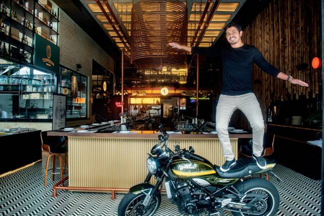 Homem de calça clara e blusa preta de mangas compridas em cima de uma moto preta e amarela com os braços abertos dentro de um espaço interno de restaurante com paredes feitas de bambu e
