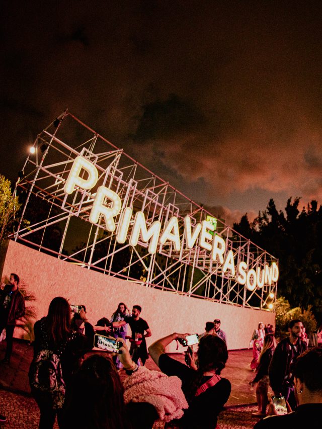 Imagem mostra outdoor escrito "Primavera Sound" com luzes iluminadas. Ao redor, pessoas transitando