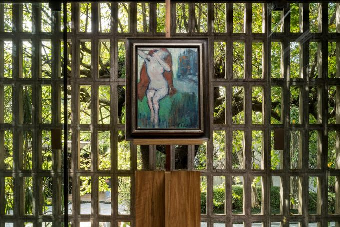Quadro de Picasso em um cavalete de madeira. Tela mostra mulher nua sentada em uma espécie de manta lilás. Ela ergue os braços dobrados. Ao fundo, é possível ver uma parede com orifícios que mostram um jardim