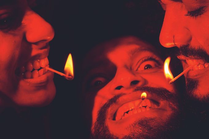Três atores negros com os rostos focados e iluminados por uma luz vermelha. Seguram palitos de fósforo acesos na boca