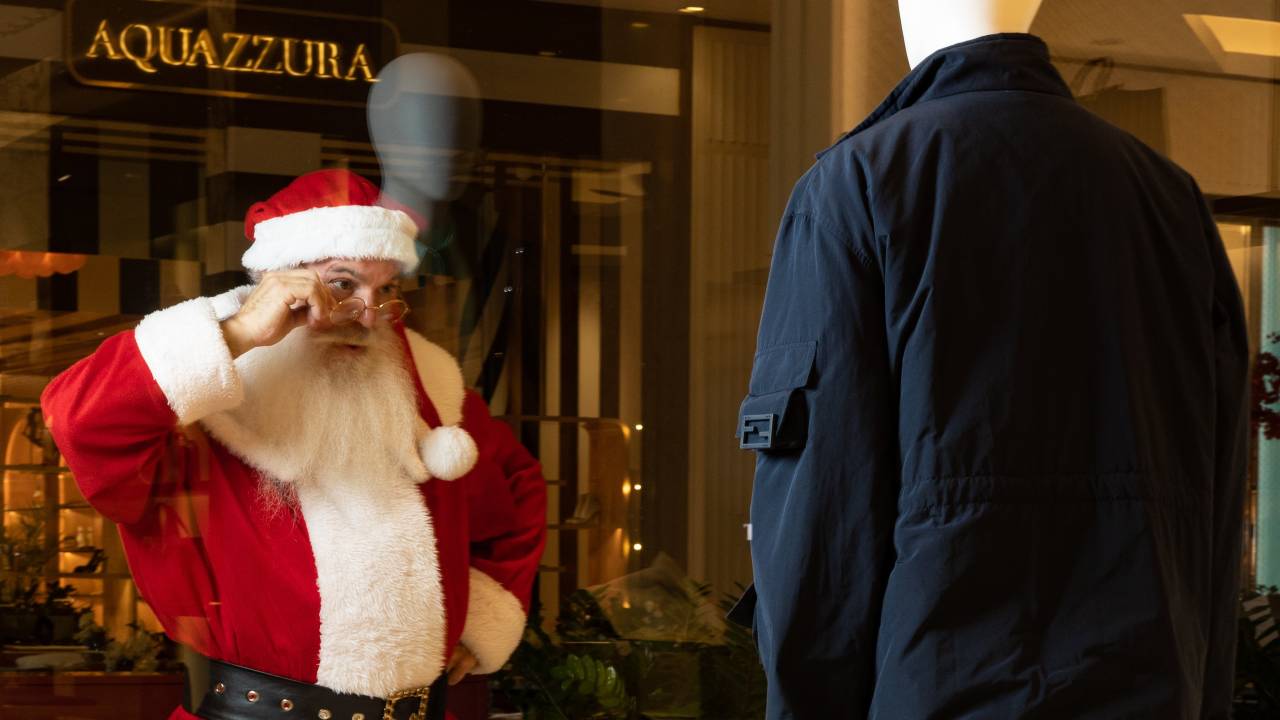 Papai Noel para em frente a vitrine e abaixa os óculos de grau para encarar manequim vestido com jaqueta azul escuro.