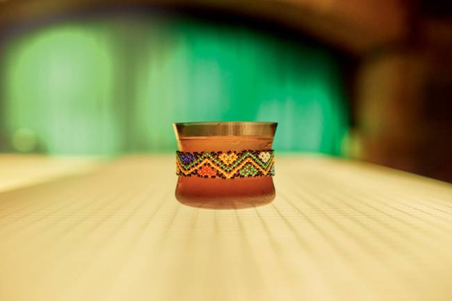 Copo baixo com borda superior dourada e uma espécie de cinta feita de miçangas coloridas formando um padrão que remete a adereços indígenas, com conteúdo alaranjado e apoiado sobre mesa de madeira clara