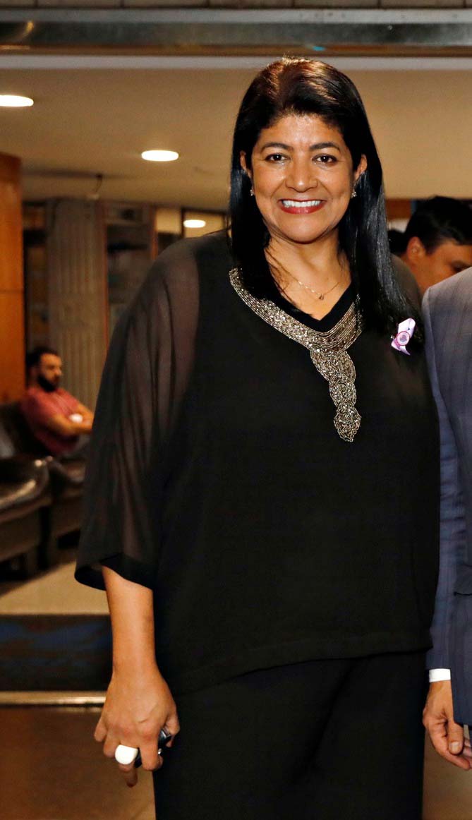 A secretária Elza Souza, uma mulher negra, de cabelos lisos, curtos e escuros. Está em pé, usa uma blusa preta e sorri para a foto