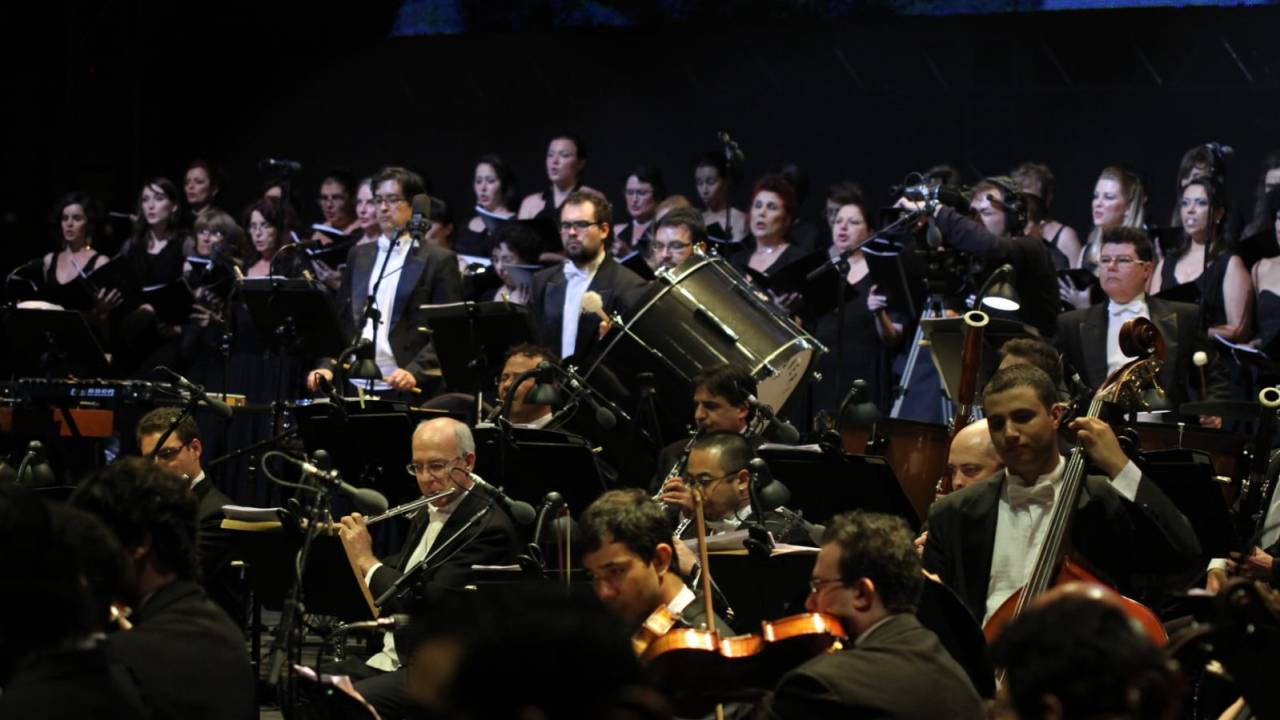 Imagem mostra orquestra com diversos homens de terno tocando instrumentos
