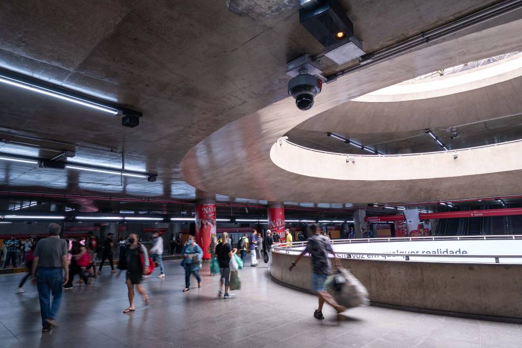 AlexandreMoreira53 Foto mostra estação da Sé. Pessoas andam e, no teto, é possível ver uma grande câmera de reconhecimento facial