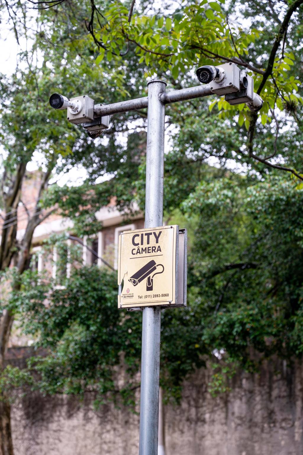 Poste com duas câmeras de monitoramento, uma em cada extremidade. Abaixo há uma placa amarela onde se lê "City Câmera"