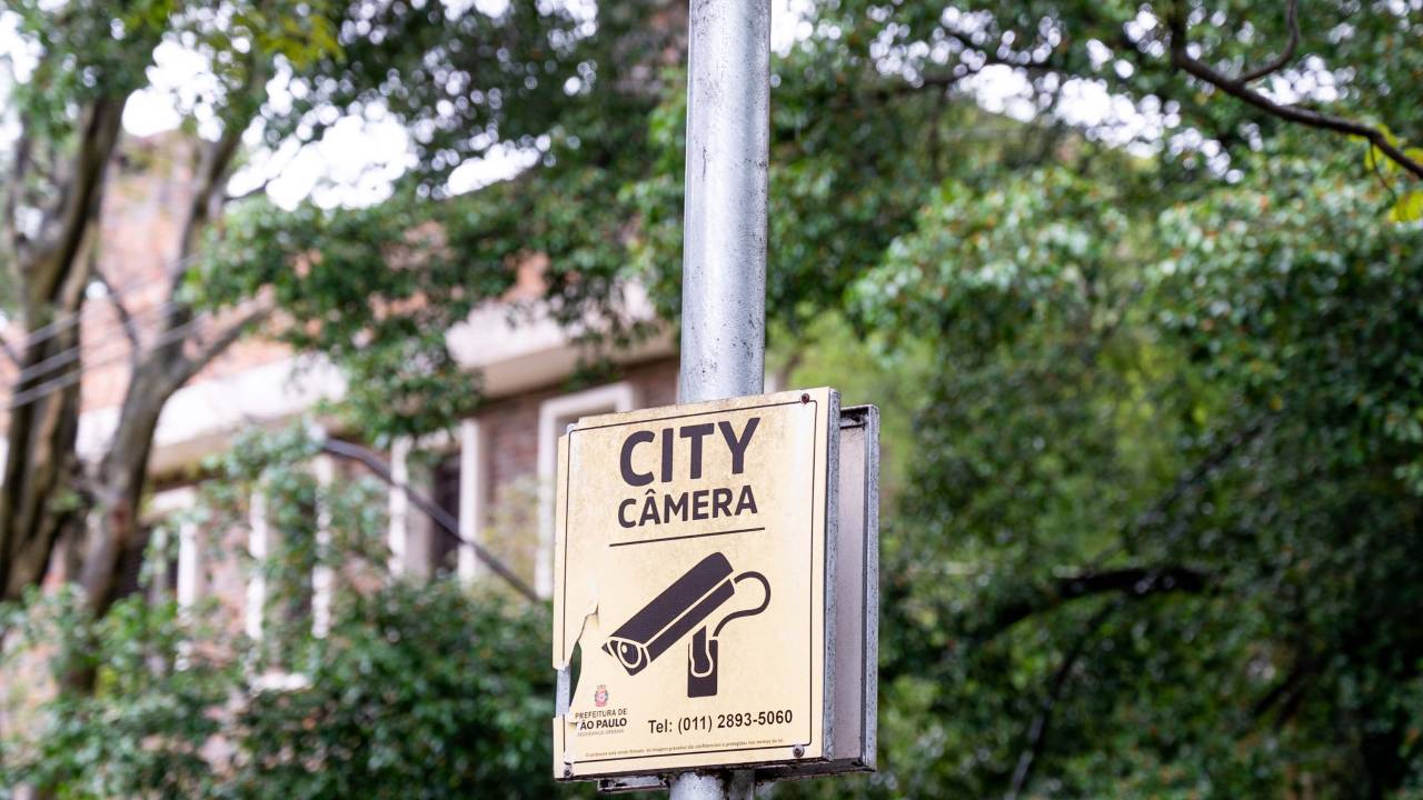 Poste com duas câmeras de monitoramento, uma em cada extremidade. Abaixo há uma placa amarela onde se lê "City Câmera"