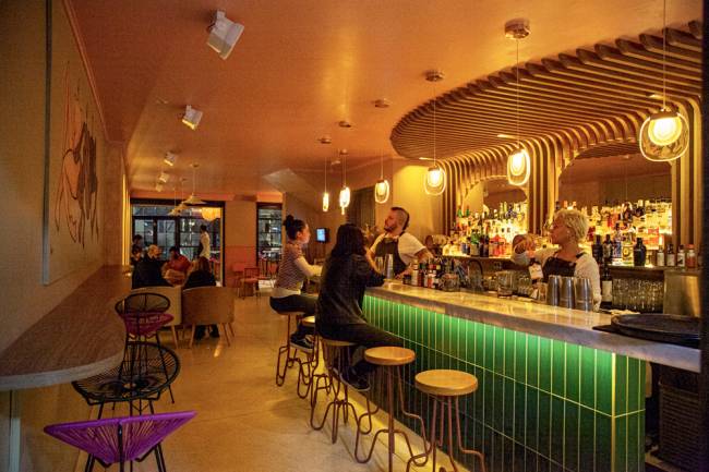 Ambiente interno de bar com luz baixa, balcão de revestimento de azulejos verdes, banquetas ao lado do balcão e tampos de madeira clara e mesas ao lado