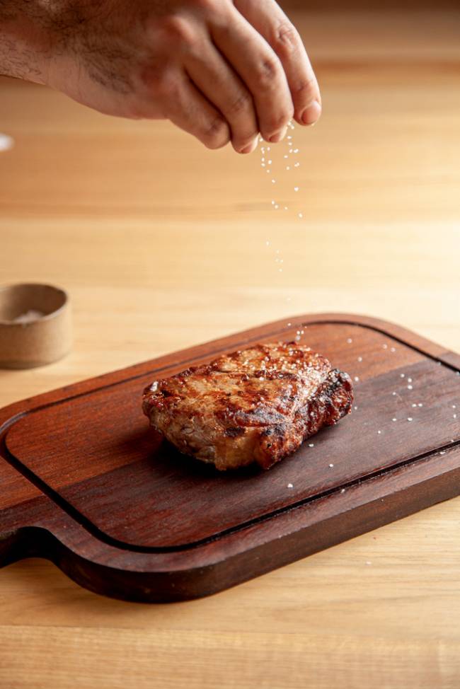 bife de chorizo apoiado sobre tábua de carne em mesa de madeira clara, com mão salpicando sal sobre a peça de carne assada