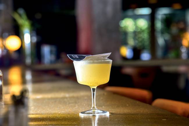 French 75 Casablanca Cineclube Cortina Taça alta com conteúdo amarelo e uma foto apoiada sobre o líquido, sobre balcão com parte de banquetas ao lado