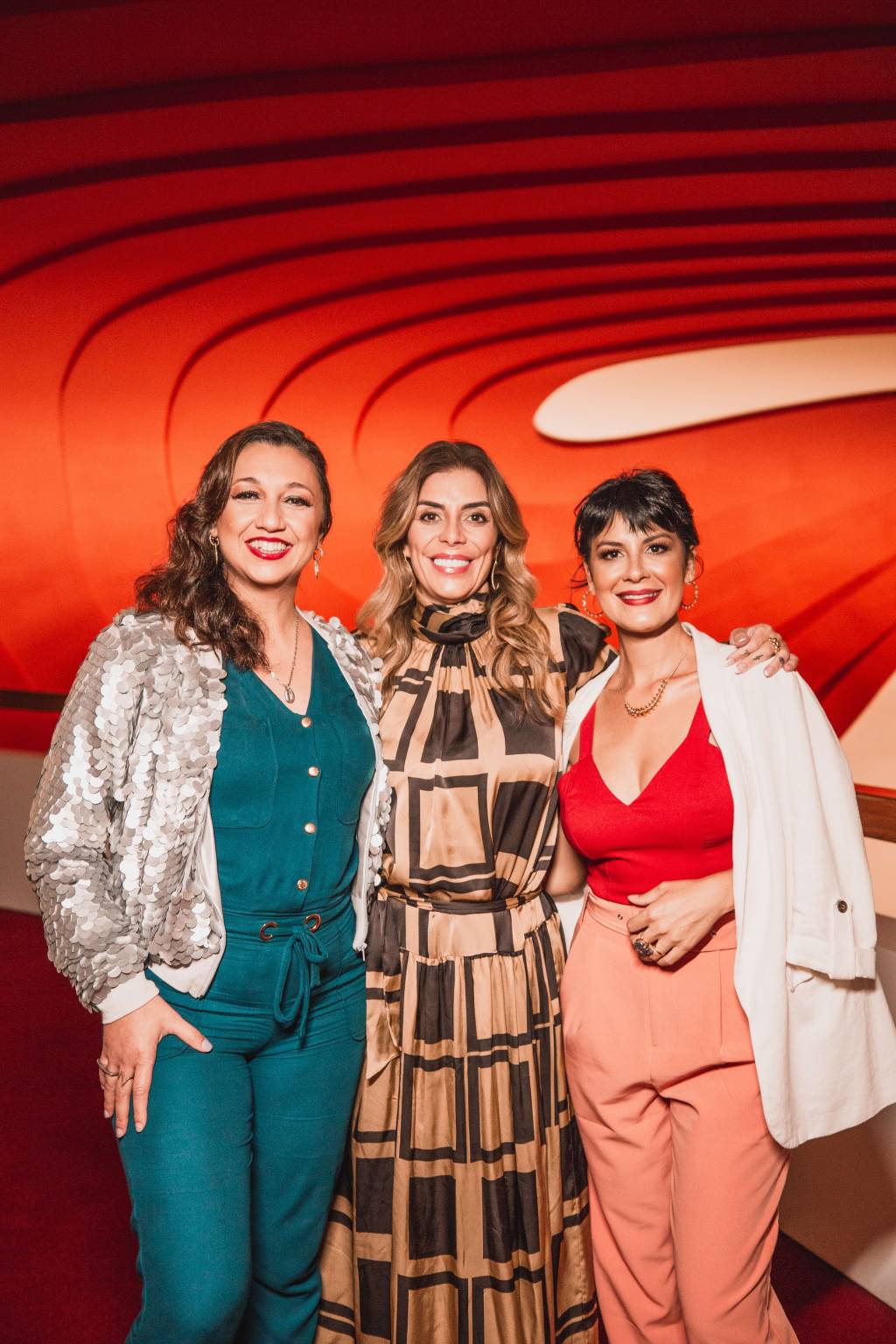 Claudia, Fatima, Monique – WME AWARDS (Mariana Smania-Divulgação) Três mulheres sorriem lado a lado. A primeira da esquerda tem cabelos longos escuros, veste blusa branca e conjunto verde. A do meio tem cabelos longos com luzes e veste conjunto estampado nas cores marrom e bege. A terceira veste blusa decotada vermelha com calça bege e mantinha branca nos ombros. Tem cabelos curtos escuros.