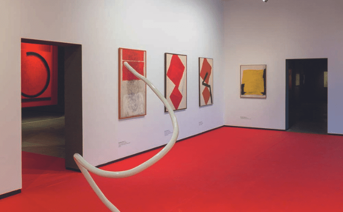 Imagem mostra espaço com paredes brancas, chão vermelho e pinturas nas paredes