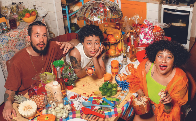 Imagem mostra três pessoas ao redor de mesa com diversas comidas e utensílios coloridos